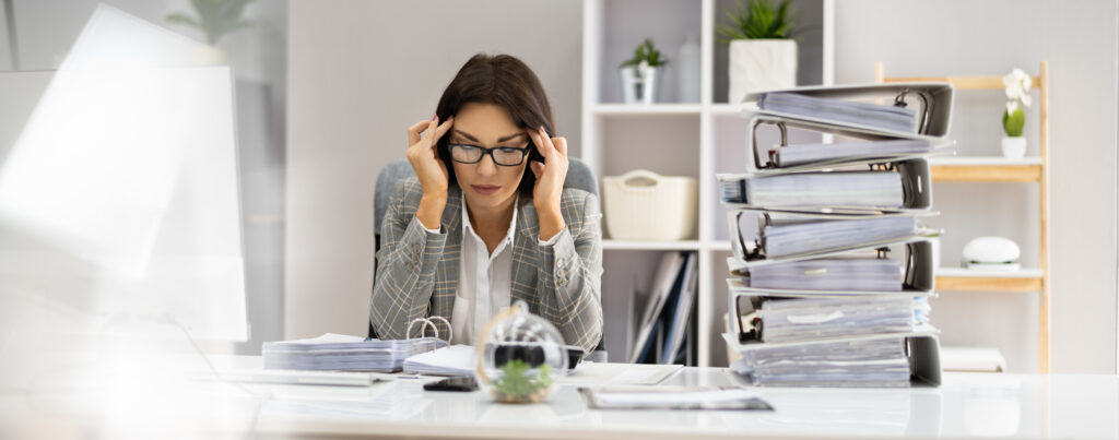Stressed office worker rubbing her temples next to a tall stack of binders.
